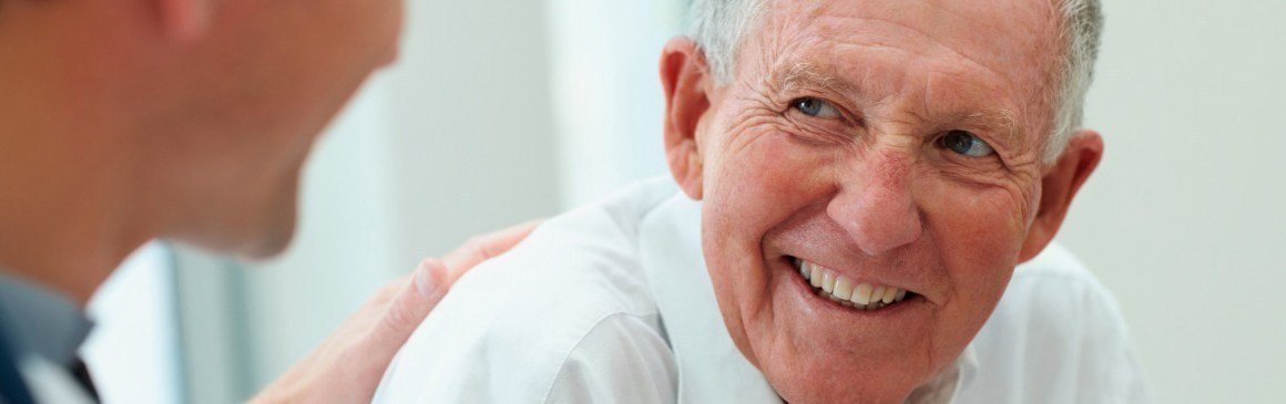 A smiling older man in conversation with a clinician, who rests a hand on his shoulder.