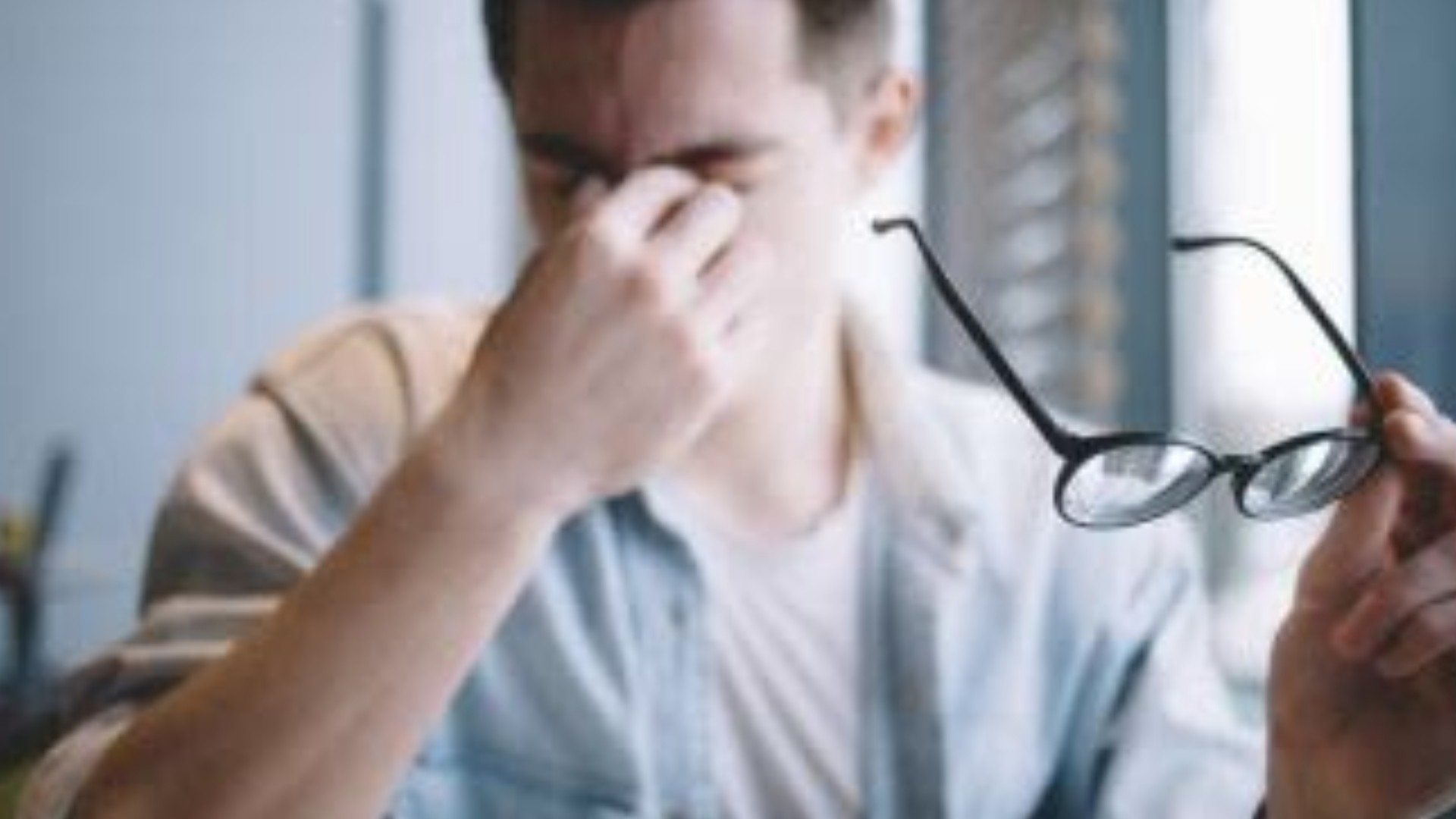 A man pinching the bridge of his nose with his eyes shut, his glasses held in the other hand.