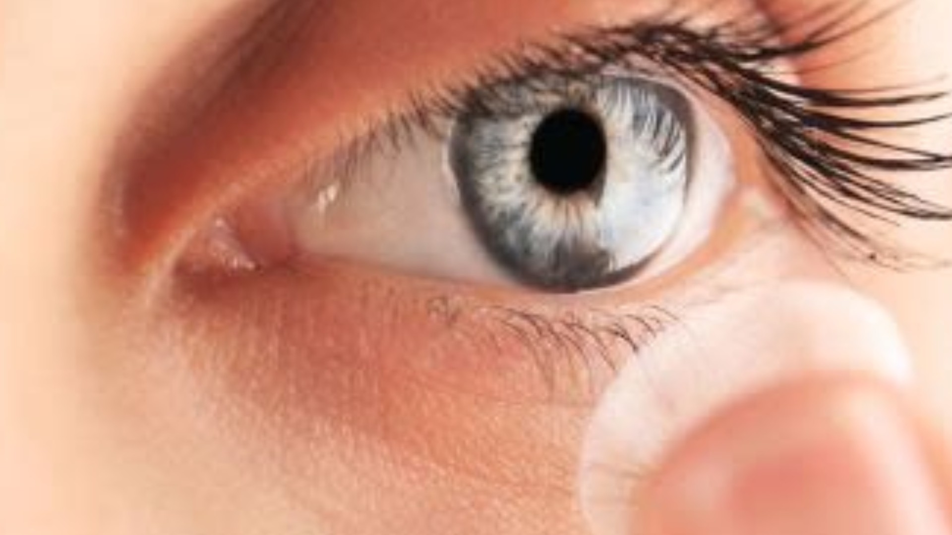 A close-up of an eye with a contact lens balanced on a fingertip in front of it.