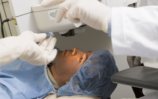 A patient in a surgical cap lies back while a gloved surgeon positions equipment above their eye.
