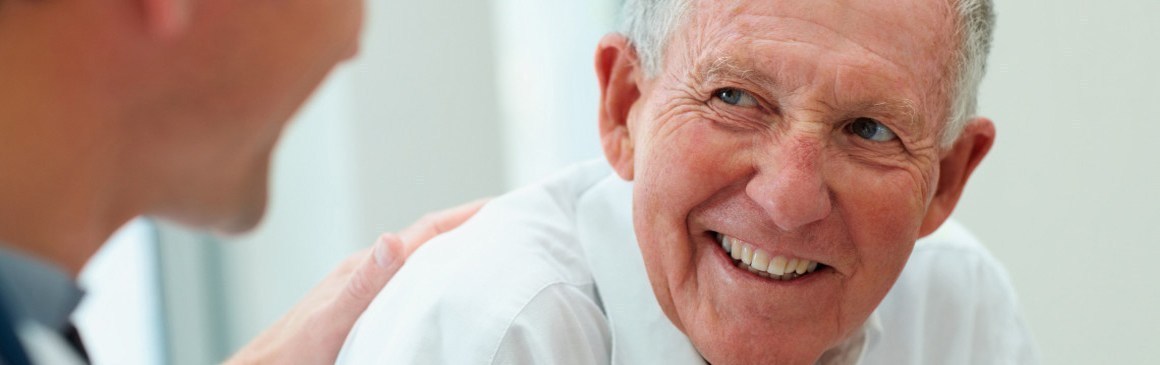 A smiling older man in conversation with a clinician, who rests a hand on his shoulder.